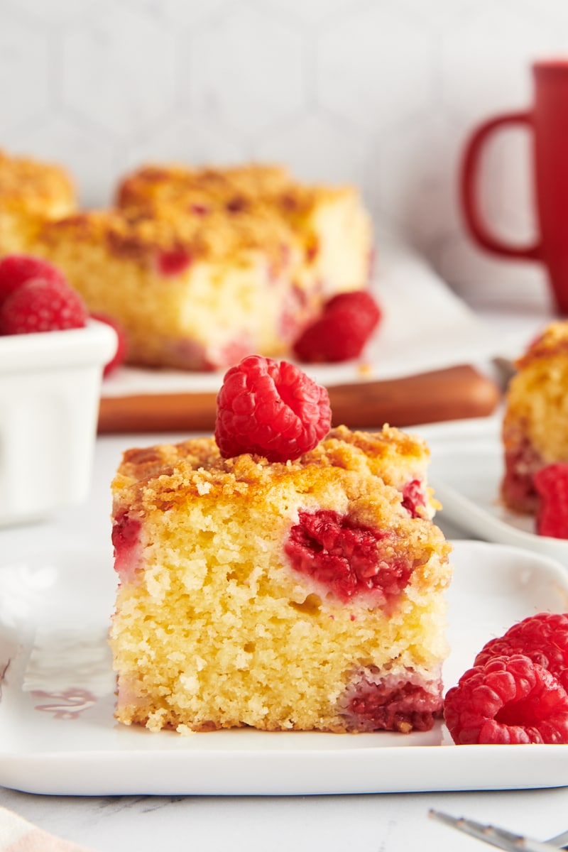 a slice of raspberry coffee cake on a white plate with more cake and a basket of raspberries in the background