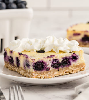 Blueberry oat cheesecake bar on white plate with pint of blueberries in background