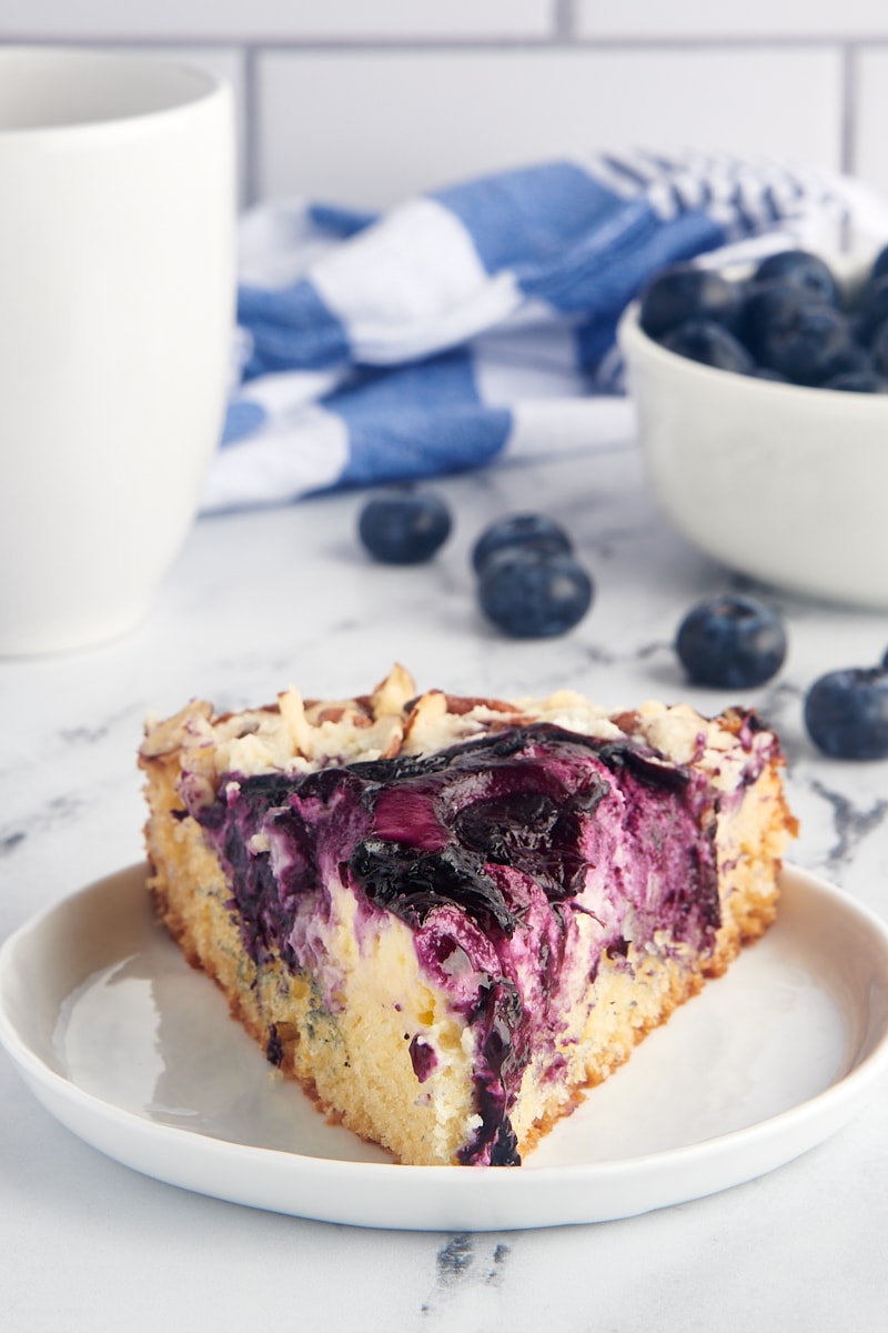 Slice of blueberry cream cheese coffee cake on white plate with blueberries in background