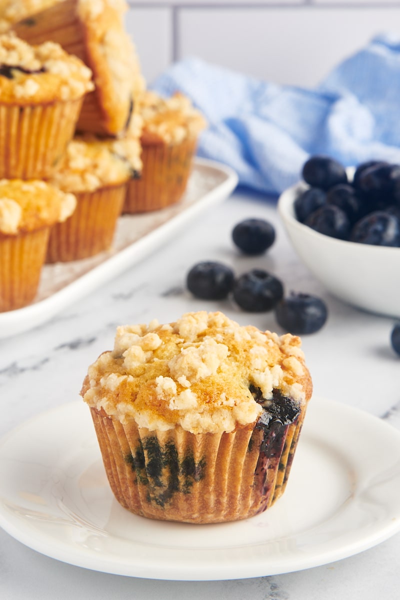 Blueberry crumb muffin on white plate with berries and additional muffins in background