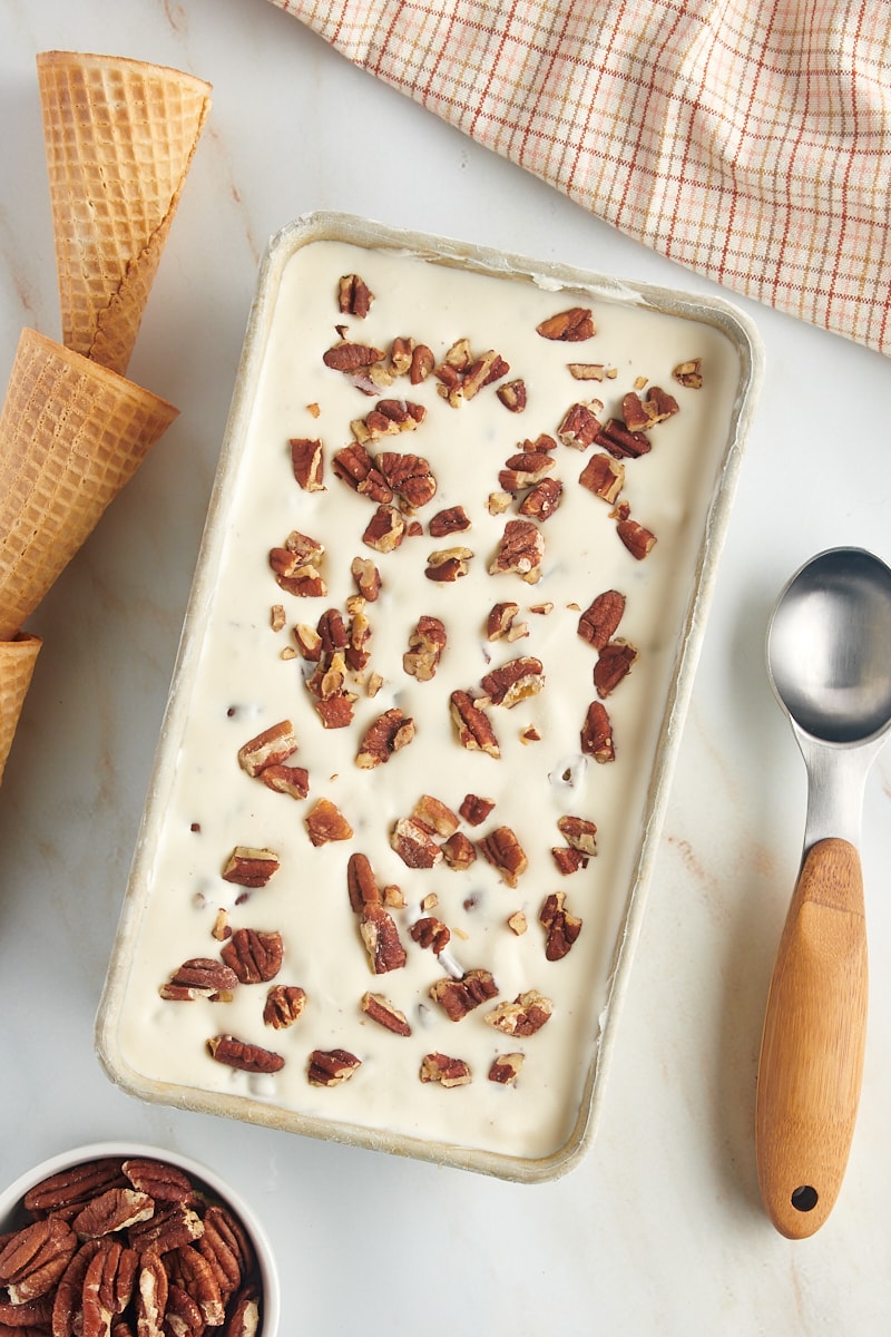 overhead view of butter pecan ice cream in a metal loaf pan with an ice cream scoop and waffle cones to the side