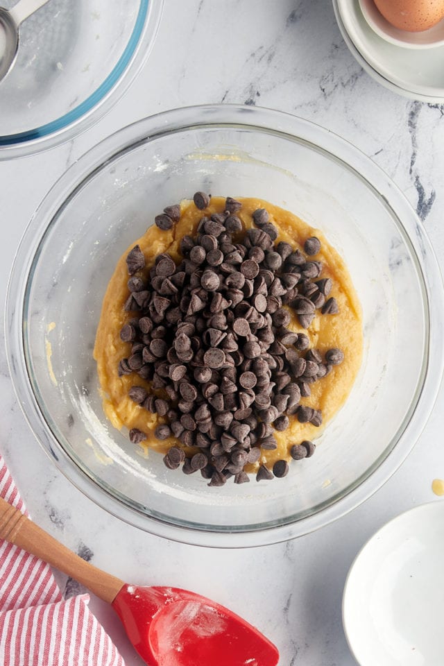 overhead view of chocolate chips added to cookie batter in a glass mixing bowl