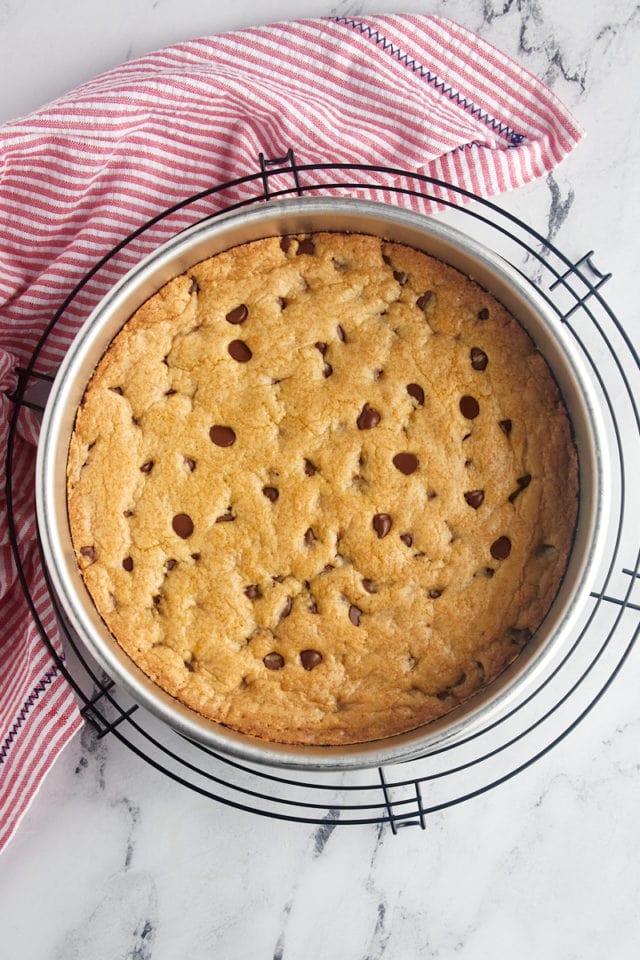 overhead view of freshly baked chocolate chip cookie cake in a cake pan on a wire rack