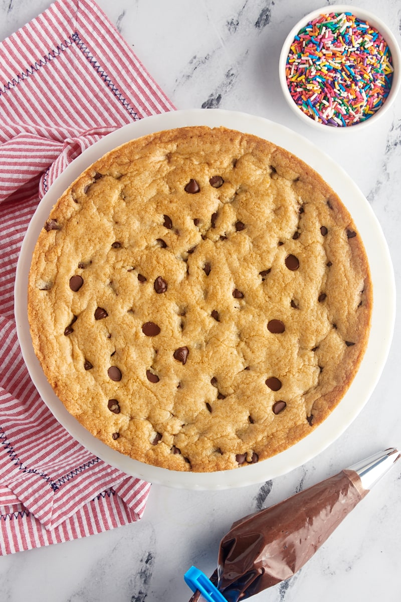 overhead view of chocolate chip cookie cake on a white cake stand with a bowl of sprinkles and chocolate buttercream frosting in a piping bag beside