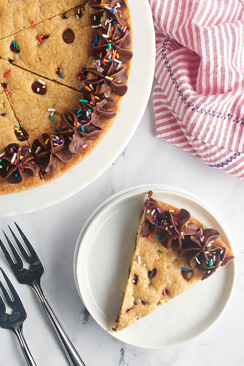overhead view of a slice of chocolate chip cookie cake on a white plate next to the remaining cookie cake on a white cake stand