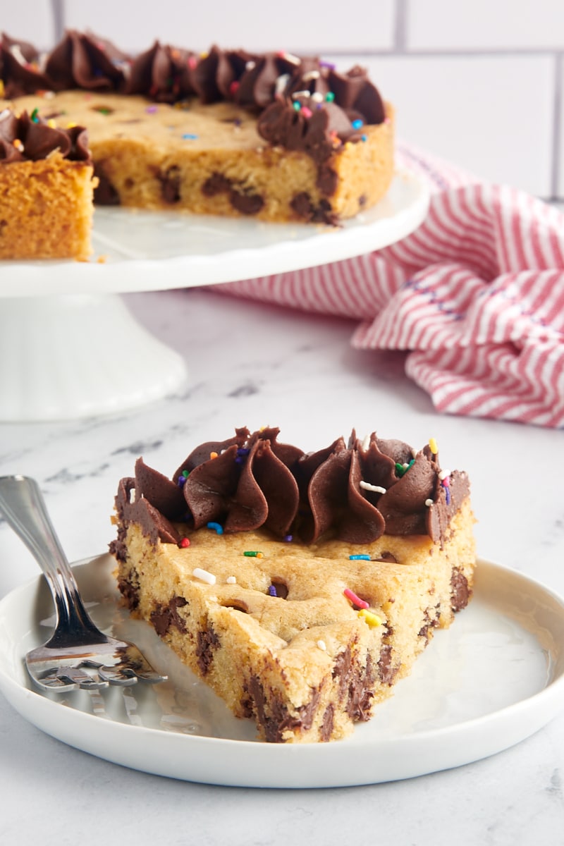 a slice of chocolate chip cookie cake on a white plate with remaining cake on a white cake stand in the background