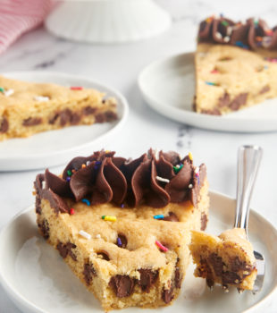 a slice of chocolate chip cookie cake on a white plate with more servings in the background