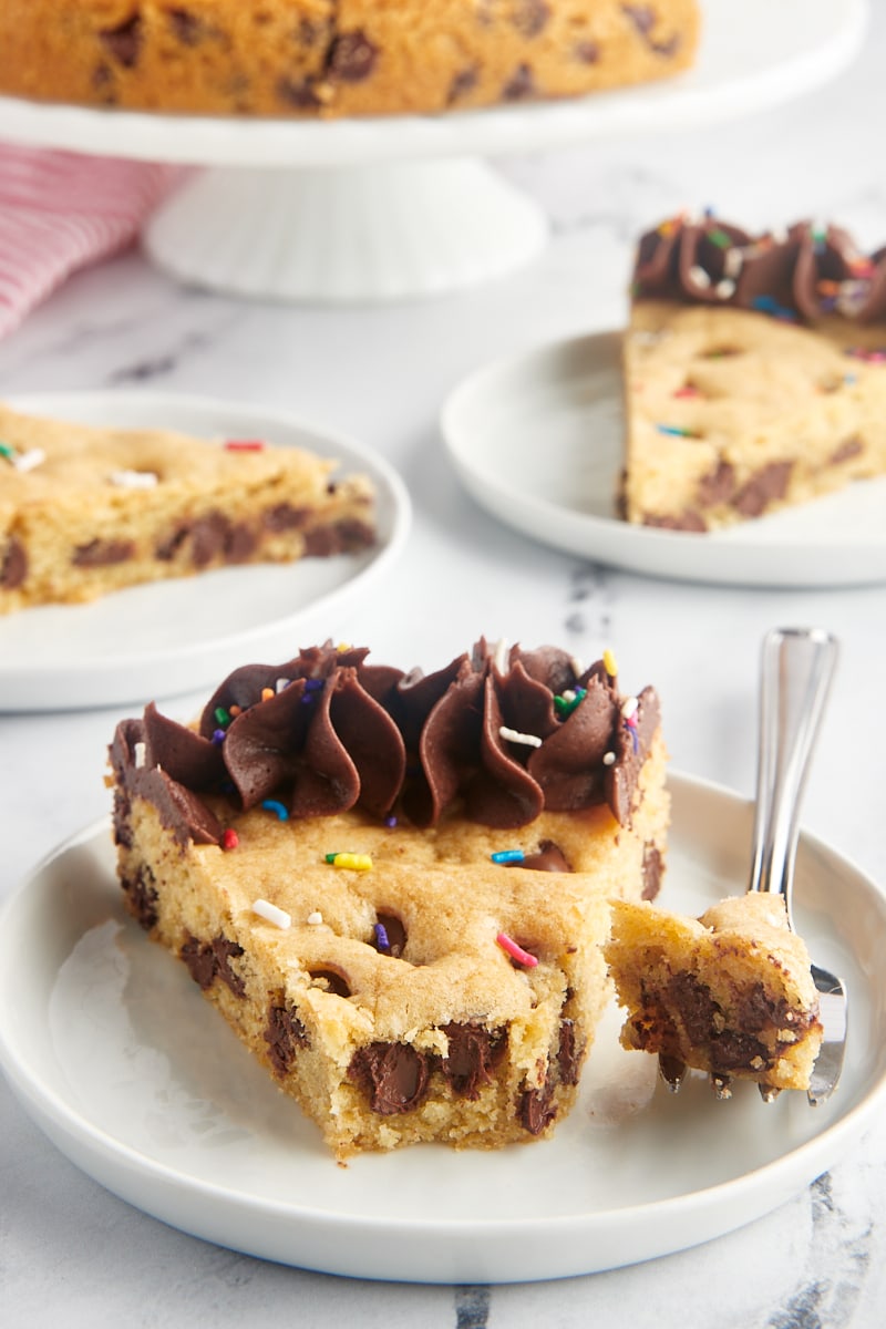 a slice of chocolate chip cookie cake on a white plate with more servings in the background