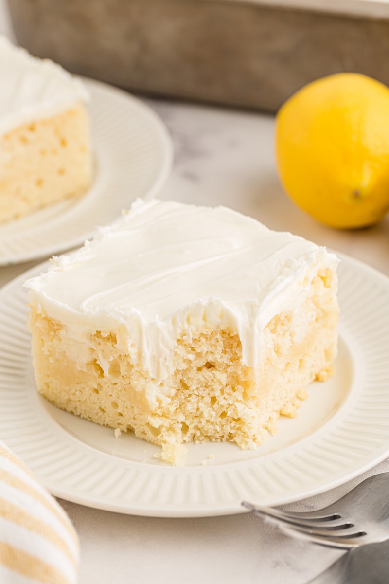 Piece of lemon poke cake on white plate, with corner eaten