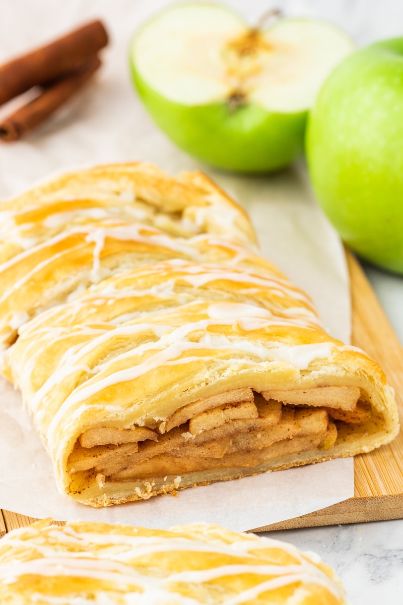 apple strudel on parchment paper on a wooden cutting board with a sliced green apple and cinnamon sticks in the background