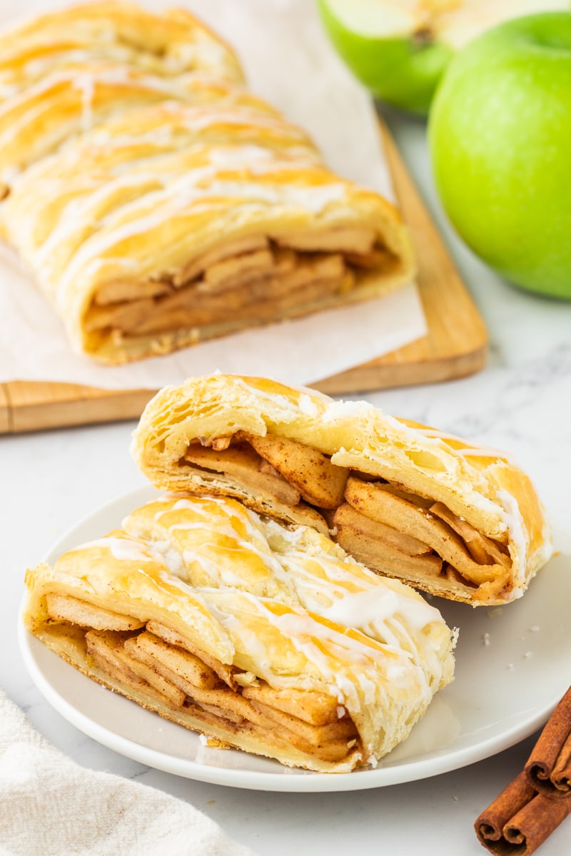 two slices of apple strudel on a white plate with more strudel and apples in the background