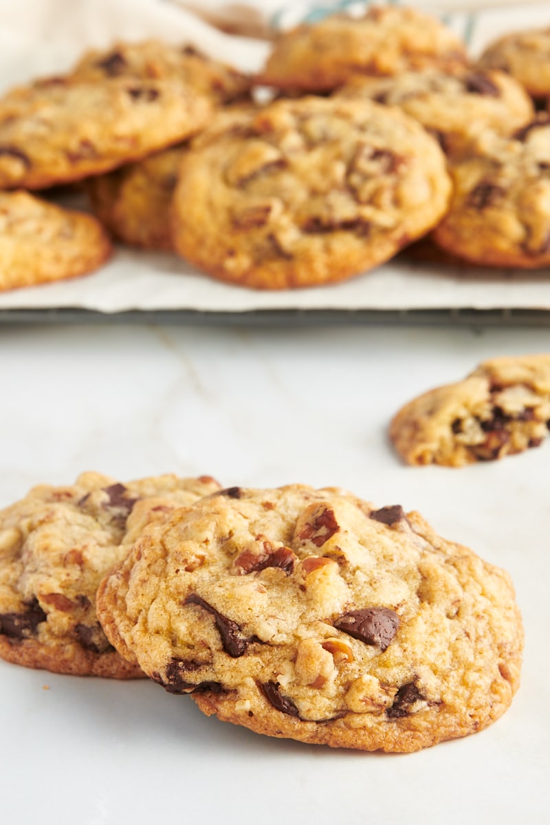 Two chocolate chunk pecan cookies stacked on countertop with additional cookies on wire rack in background