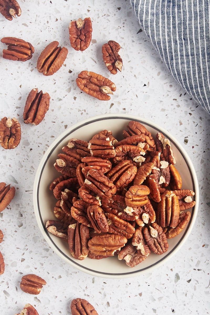 overhead view of toasted pecans in a shallow bowl with more pecans scattered around them