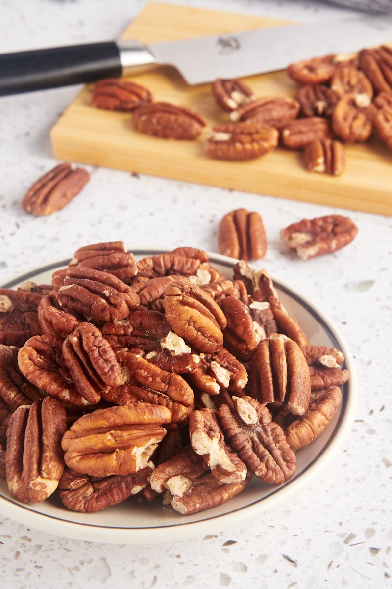 a bowl of toasted pecans with more pecans on a cutting board in the background