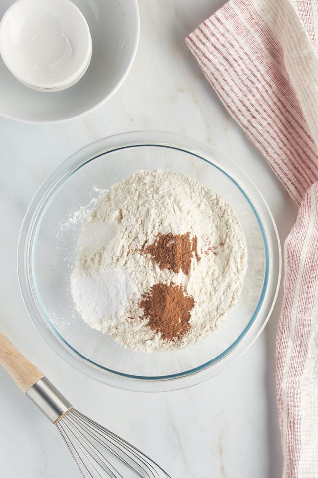 Overhead view of flour, cinnamon, nutmeg, baking soda, and salt in a glass mixing bowl.