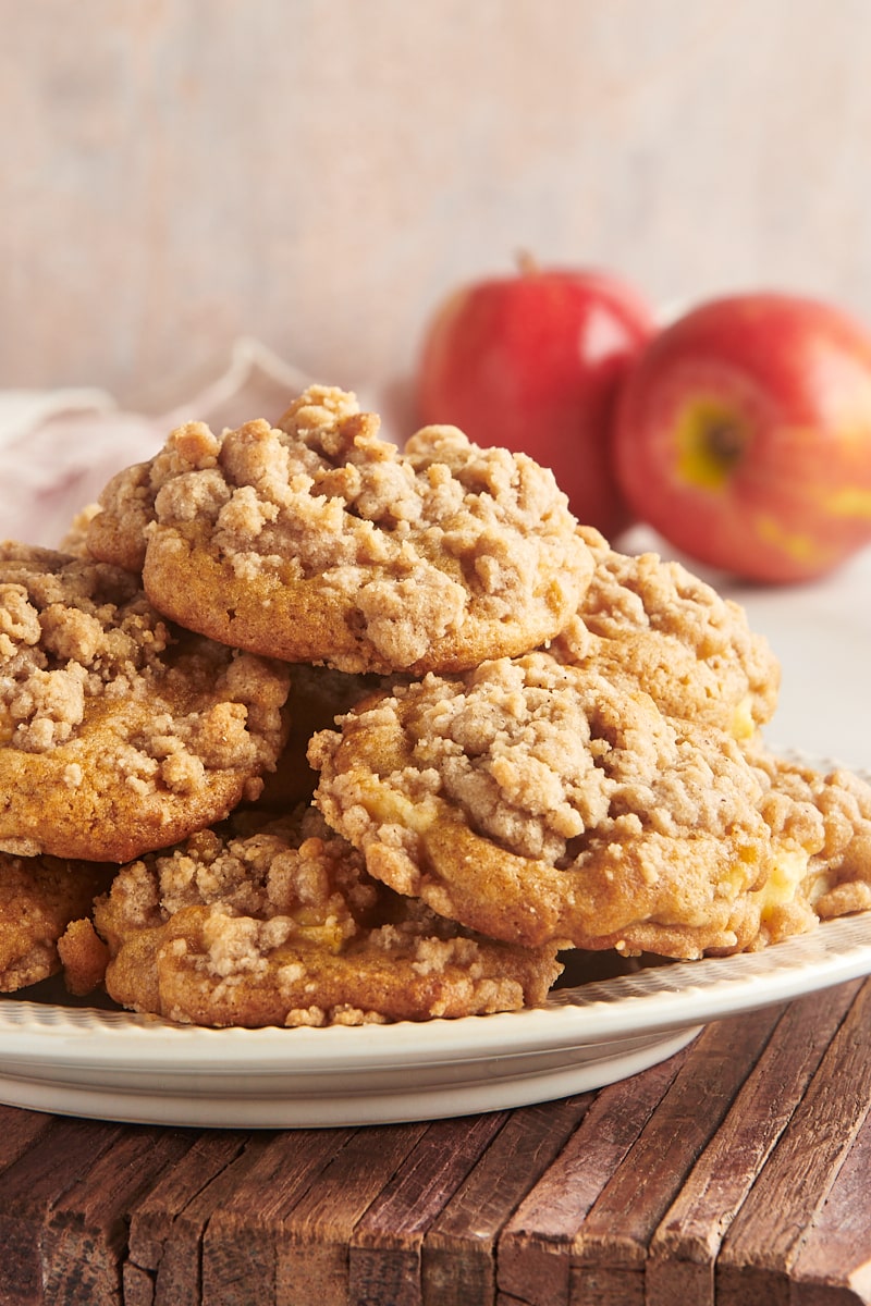 crumb-topped apple cookies piled on a plate with two apples in the background