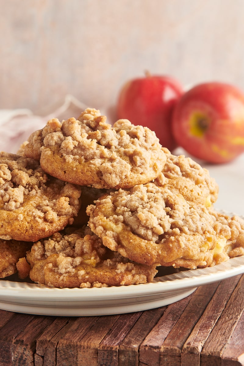 Crumb-topped apple cookies piled on a plate with two apples in the background.