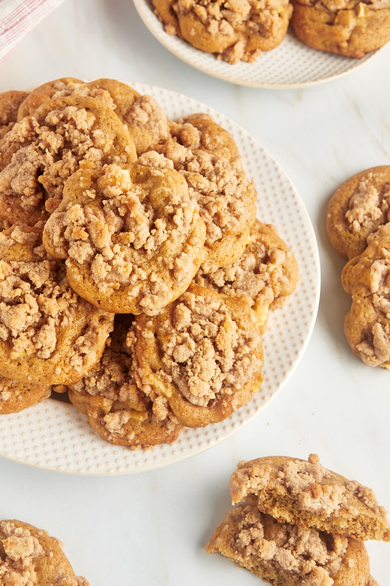 Crumb-topped apple cookies piled on a plate and scattered on a marble countertop.