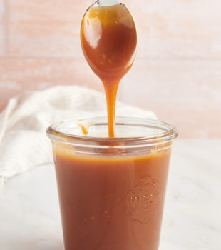 a glass jar of caramel sauce with a spoon over it with caramel dripping off of it