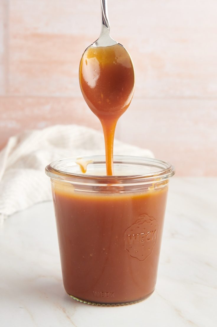a glass jar of caramel sauce with a spoon over it with caramel dripping off of it