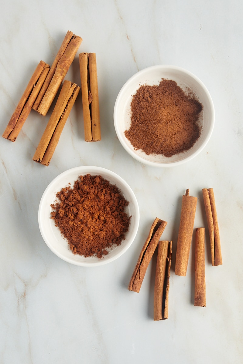 overhead view of ground cinnamon in white bowls and cinnamon sticks on a marble countertop