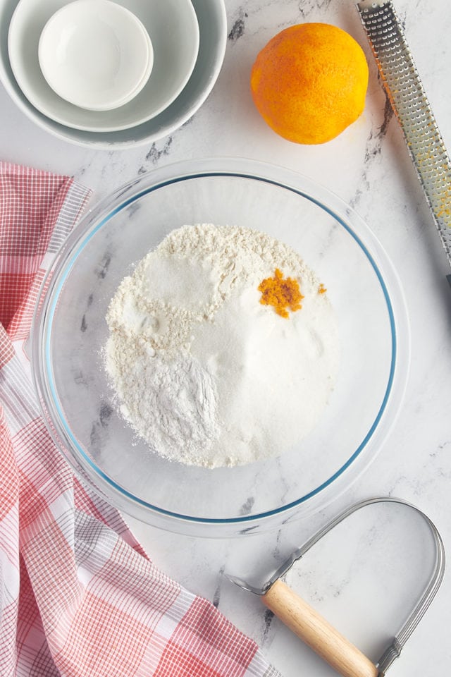 overhead view of flour, sugar, orange zest, baking powder, baking soda, and salt in a glass mixing bowl