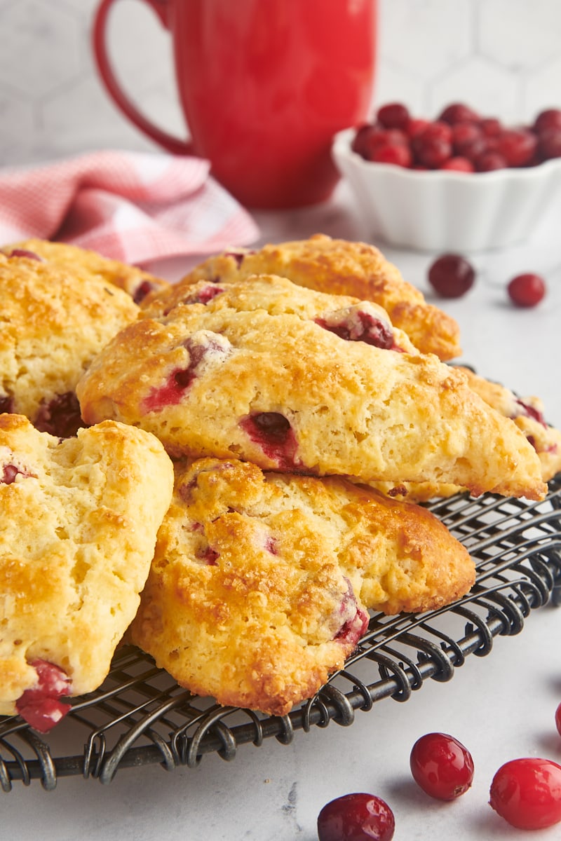cranberry orange scones on a wire rack with a bowl of cranberries and a red mug in the background
