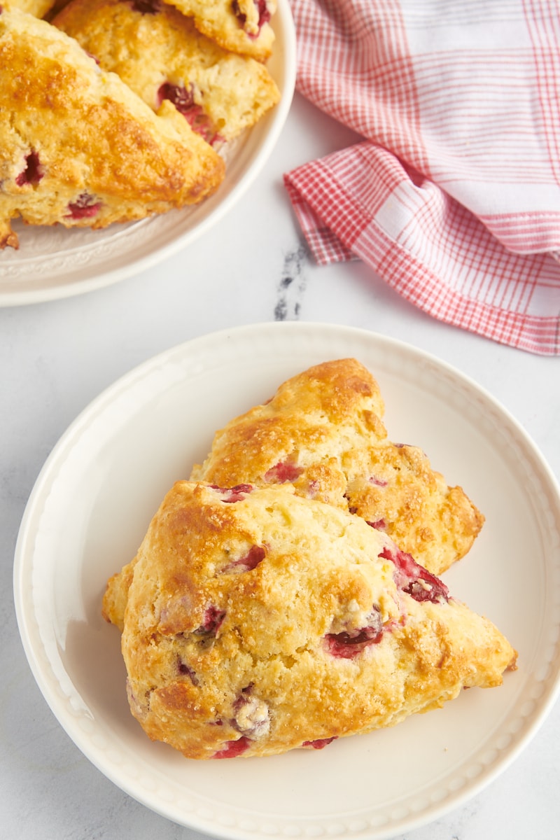 two cranberry orange scones on a plate with more scones on a large plate alongside