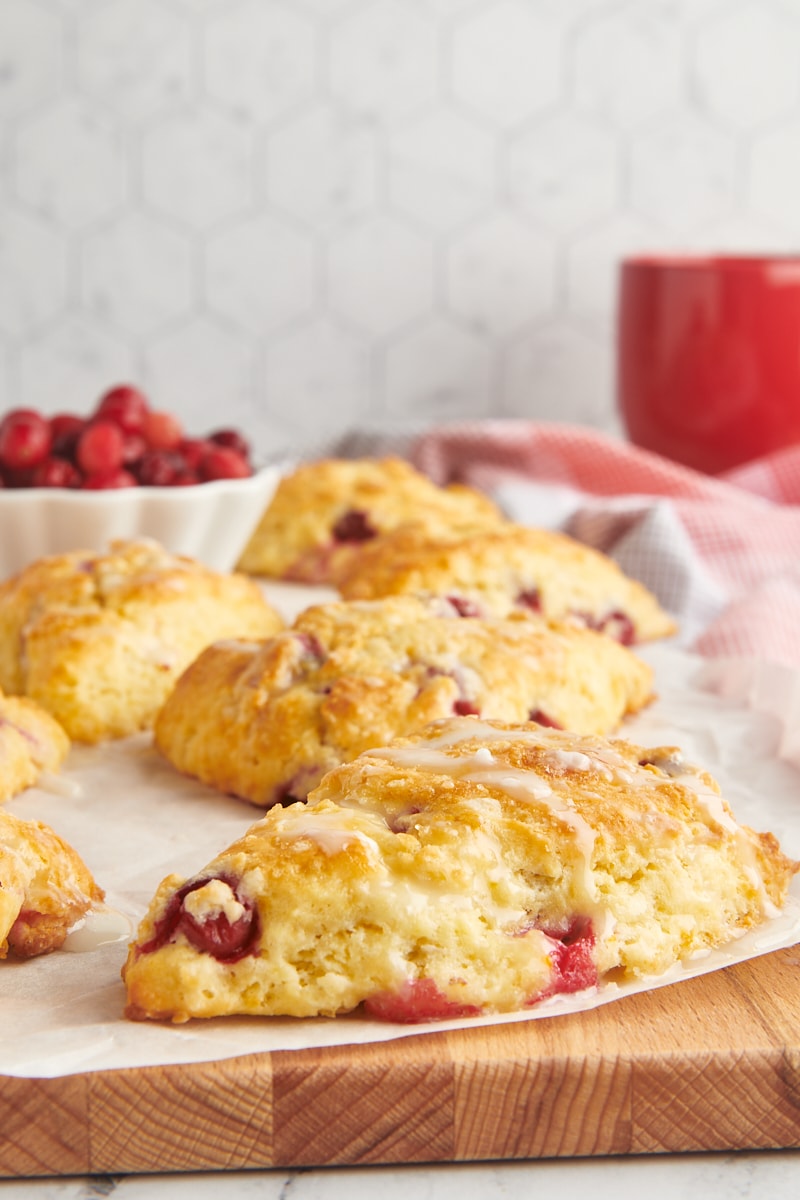 cranberry orange scones on parchment paper on top of a wooden cutting board with a bowl of cranberries and a red mug in the background