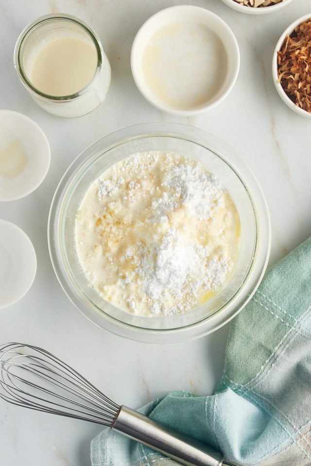 overhead view of confectioners' sugar, melted butter, vanilla extract, almond extract, and cream in a glass mixing bowl