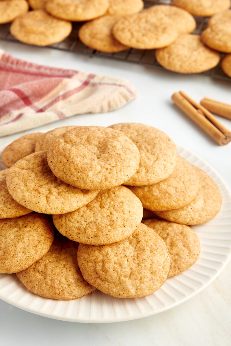 Pile of snickerdoodles on a white plate with a wire rack of cookies in the background.