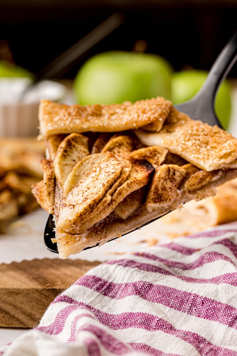 a slice of apple galette on a pie server with more pie and apples in the background