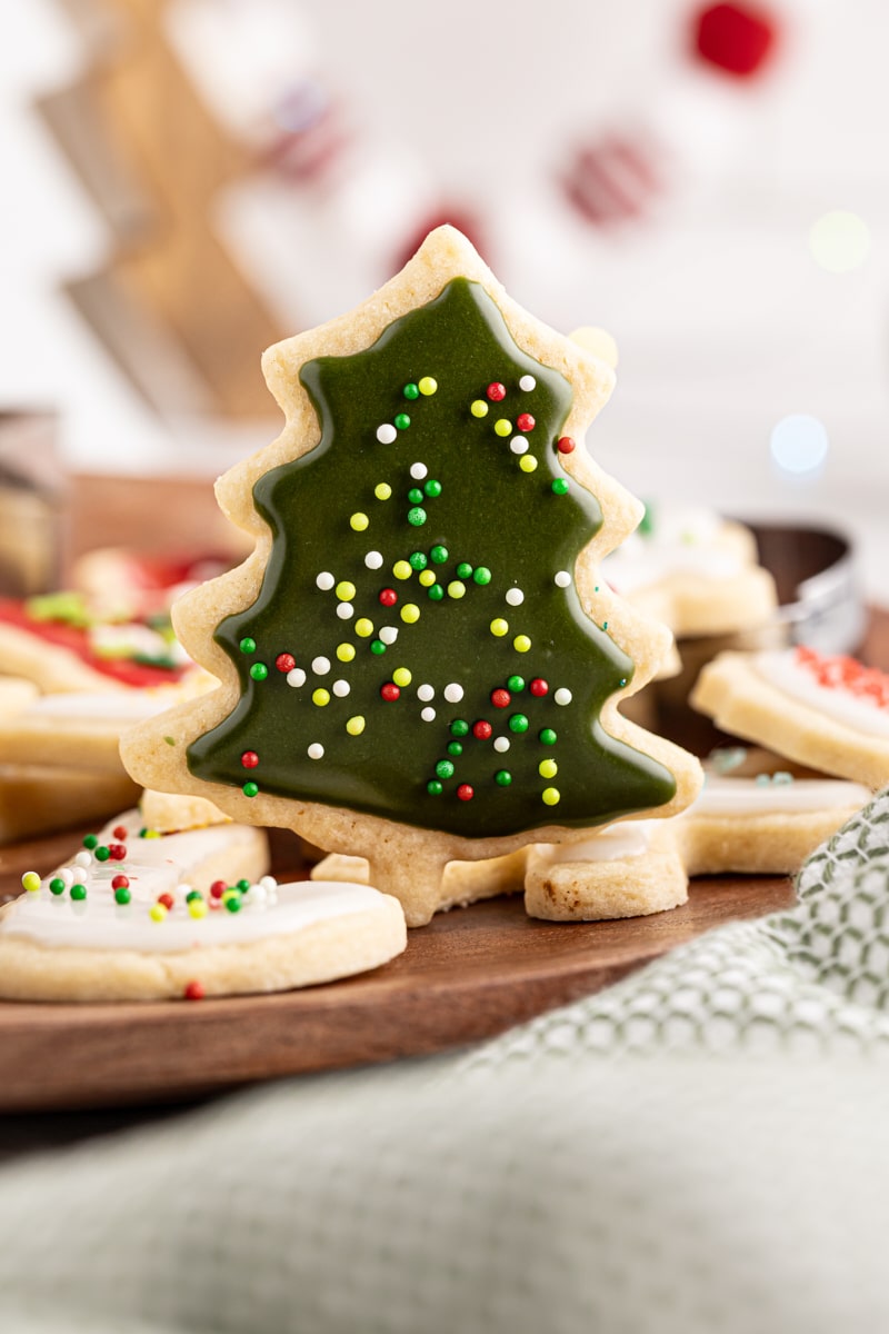 a Christmas tree-shaped cut-out sugar cookie surrounded by more cookies on a wooden tray