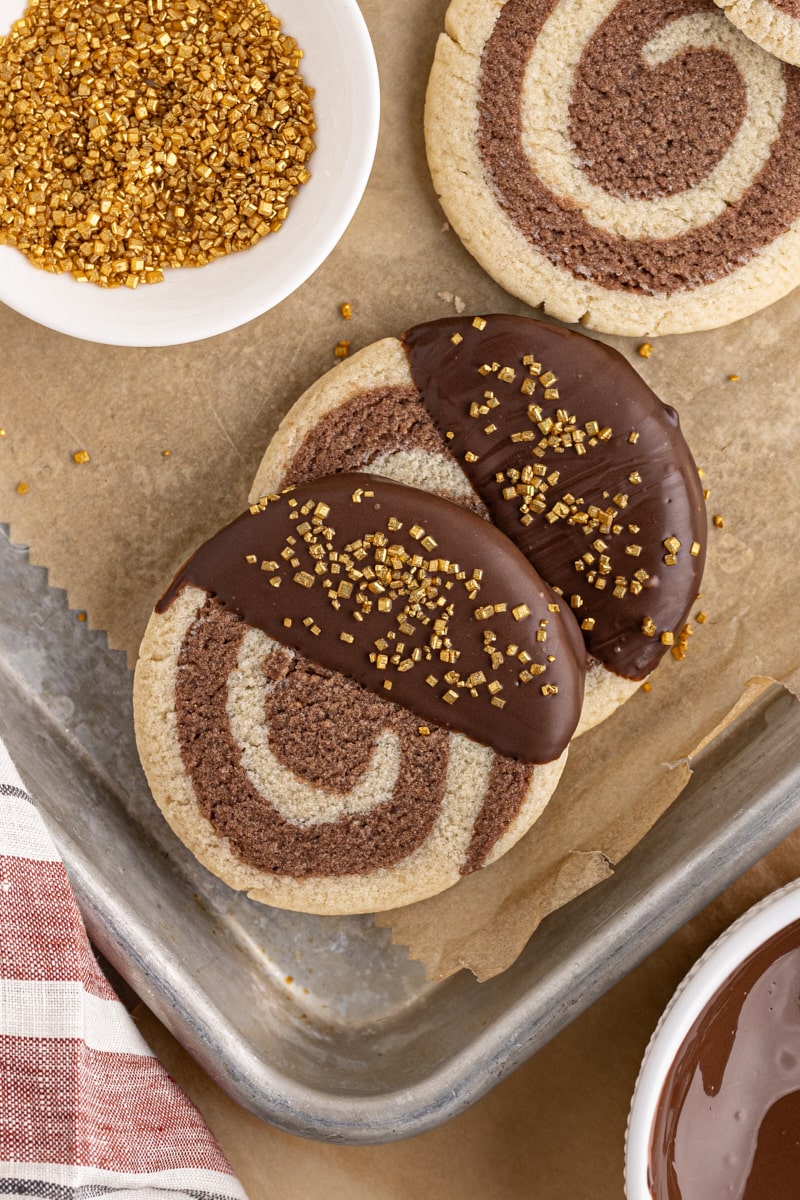 overhead view of two chocolate-dipped pinwheel cookies on a parchment- lined baking sheet with more cookies, melted chocolate, and gold sprinkles surrounding