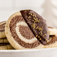 a chocolate-dipped pinwheel cookie propped against several other cookies on a white cake stand