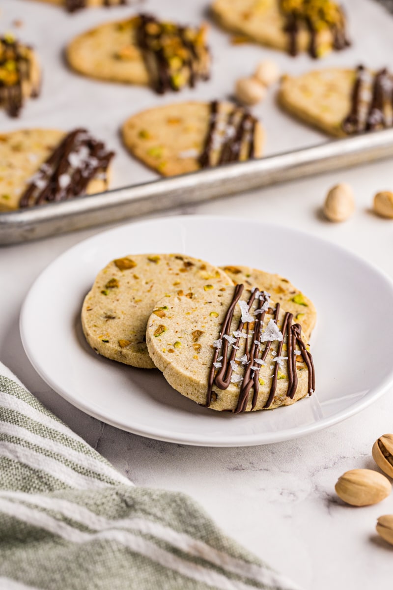 three pistachio cookies on a white plate with more cookies in the background