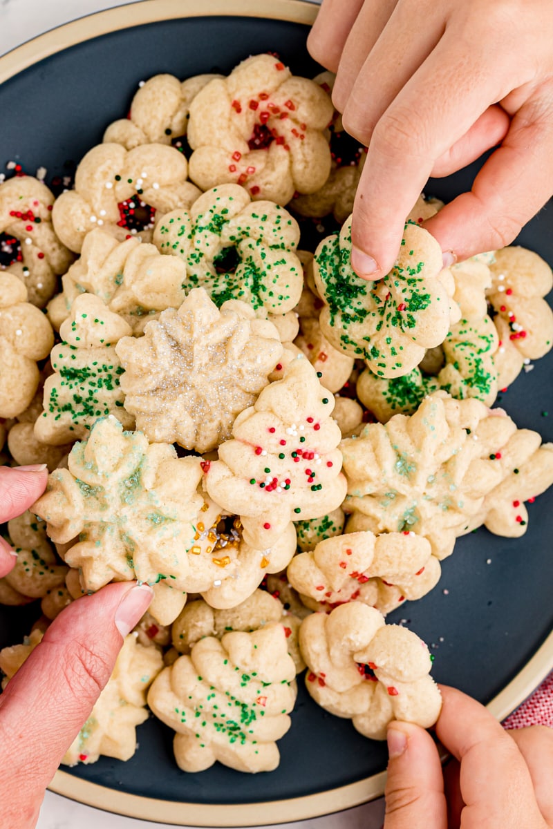 overhead view of three hands reaching for a spritz cookie from a plate full of them