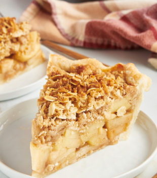 a slice of apple crumb pie on a white plate with more pie in the background