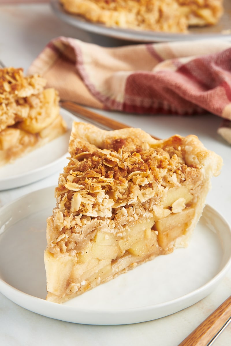 a slice of apple crumb pie on a white plate with more pie in the background