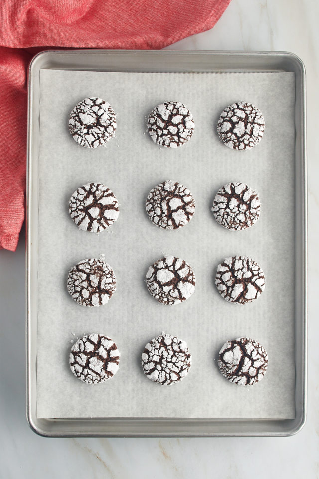 Overhead view of freshly baked chocolate crinkle cookies on a parchment-lined baking sheet.