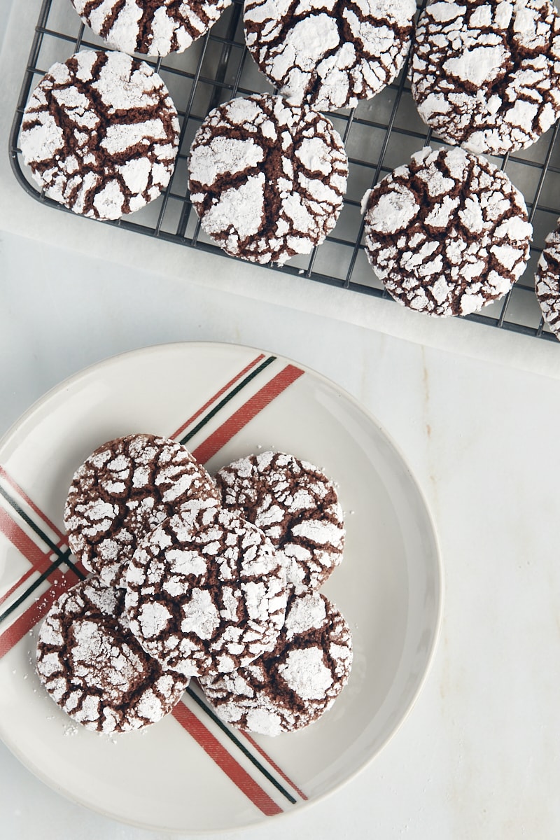 Overhead view of several chocolate crinkle cookies on a red and green striped plate with more cookies on a cooling rack beside it.
