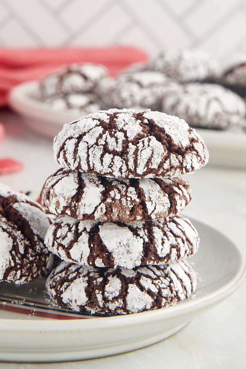 A stack of four chocolate crinkle cookies on a plate surrounded by more cookies.