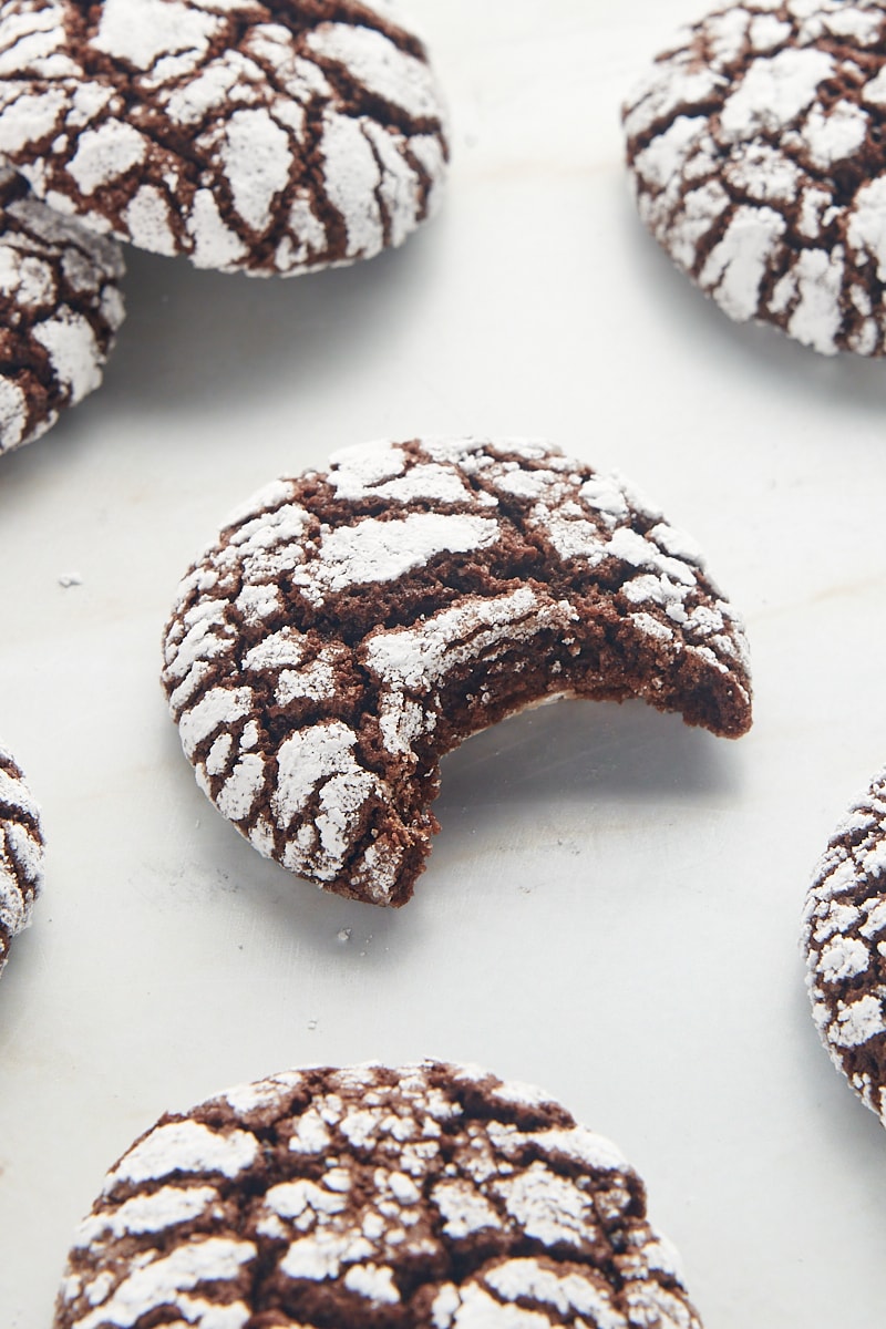 A chocolate crinkle cookie with a bite missing surrounded by more cookies on a white surface.