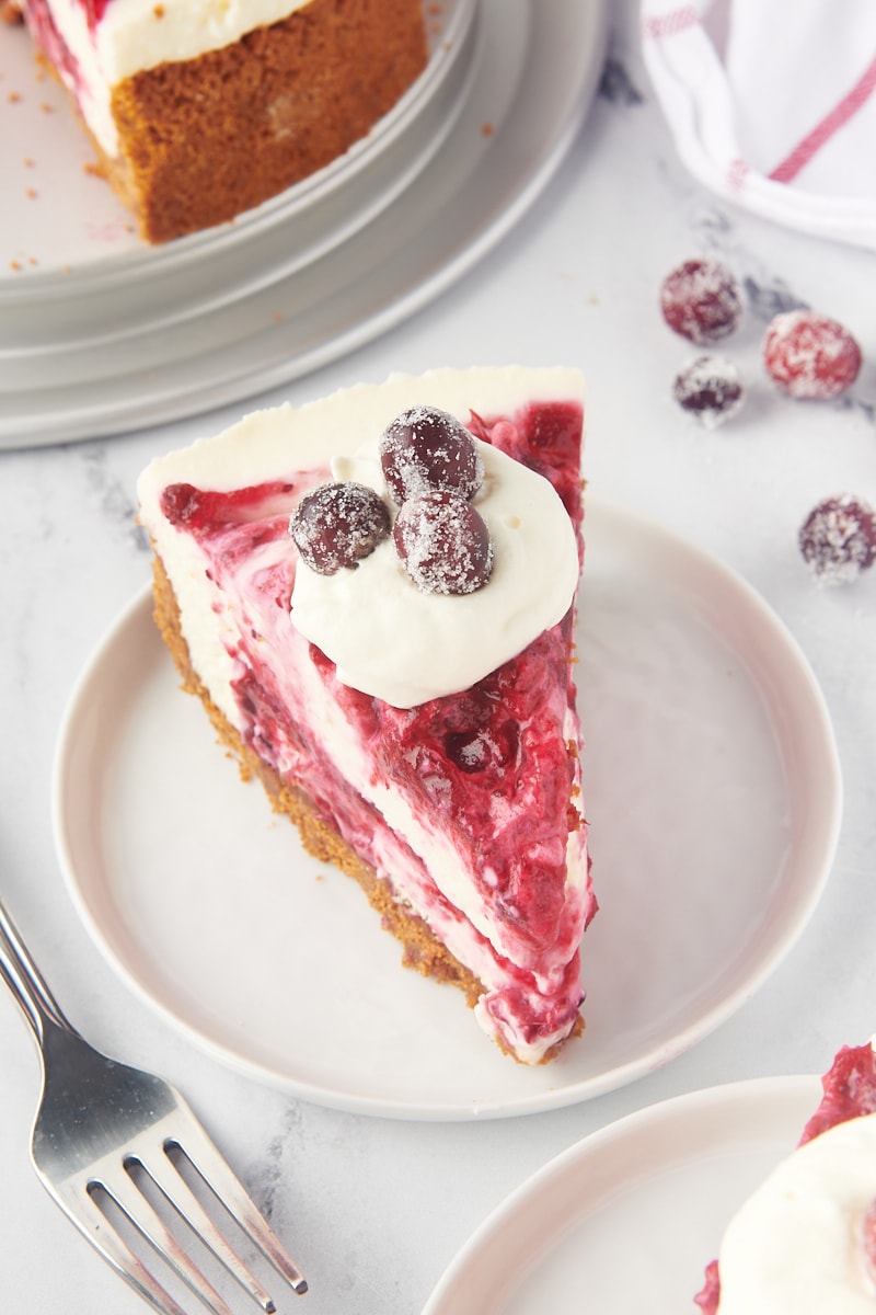 a slice of no-bake cranberry cheesecake on a white plate with more cheesecake and sugared cranberries surrounding