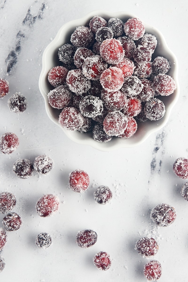 overhead view of sugared cranberries in a white bowl with more cranberries scattered around