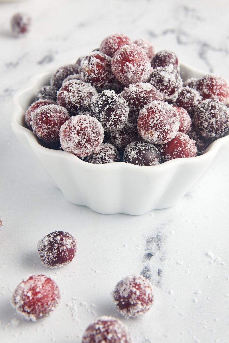 a bowl of sugared cranberries on a marble surface with more cranberries scattered around the bowl