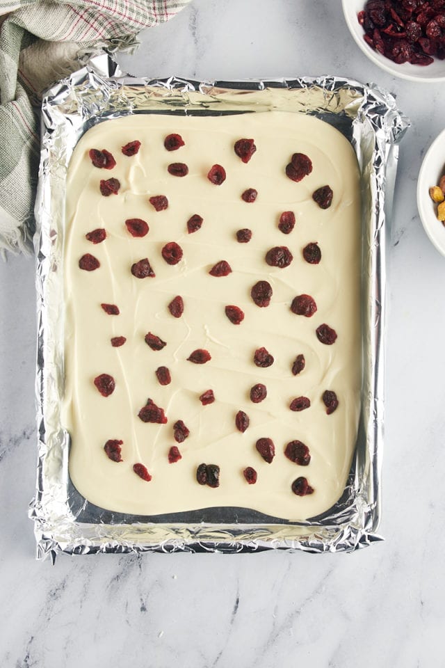 overhead view of dried cranberries scattered over melted white chocolate on a foil-lined baking sheet