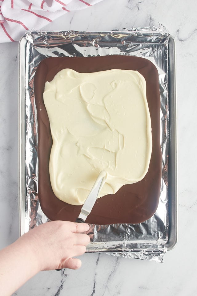 overhead view of melted white chocolate being spread over chilled dark chocolate on a foil-lined baking pan
