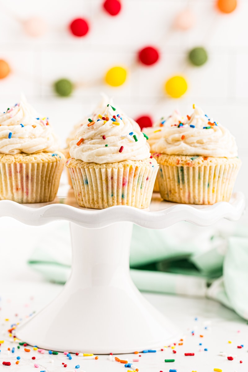 confetti cupcakes on a white cake stand with colorful garland in the background