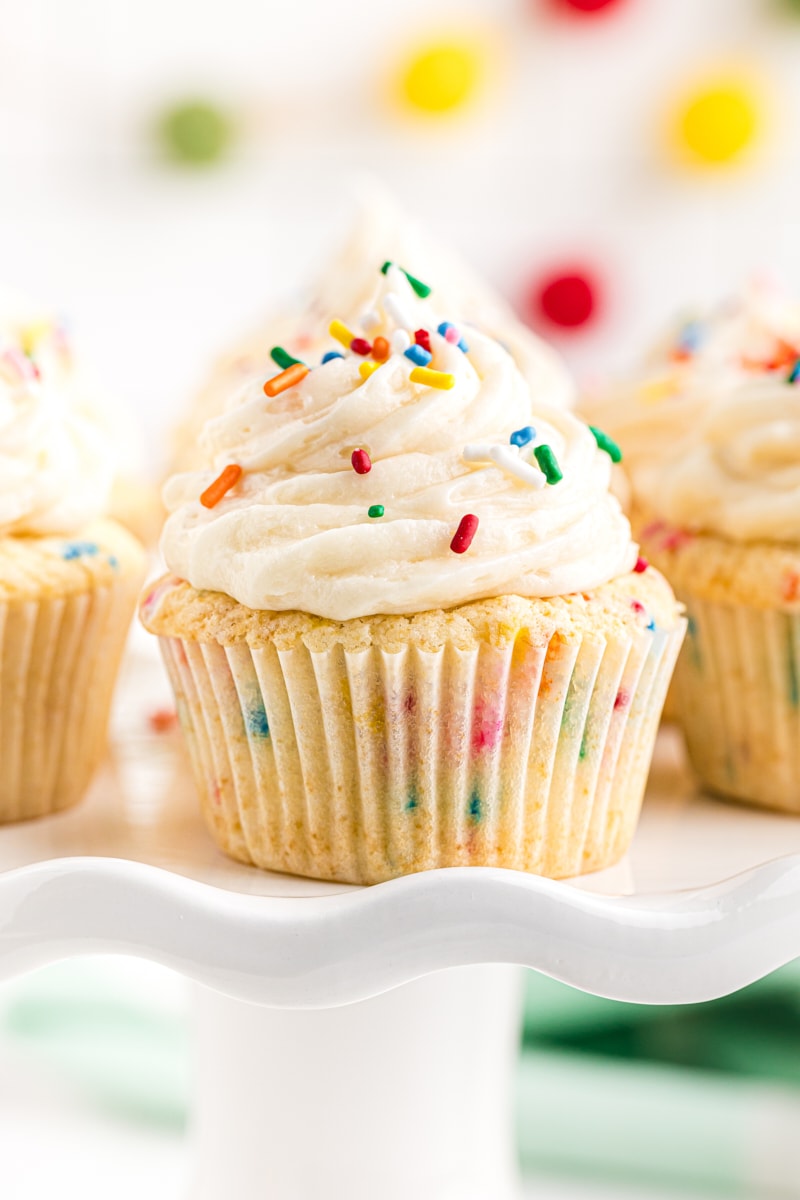a confetti cupcake on a white cake stand with more cupcakes in the background